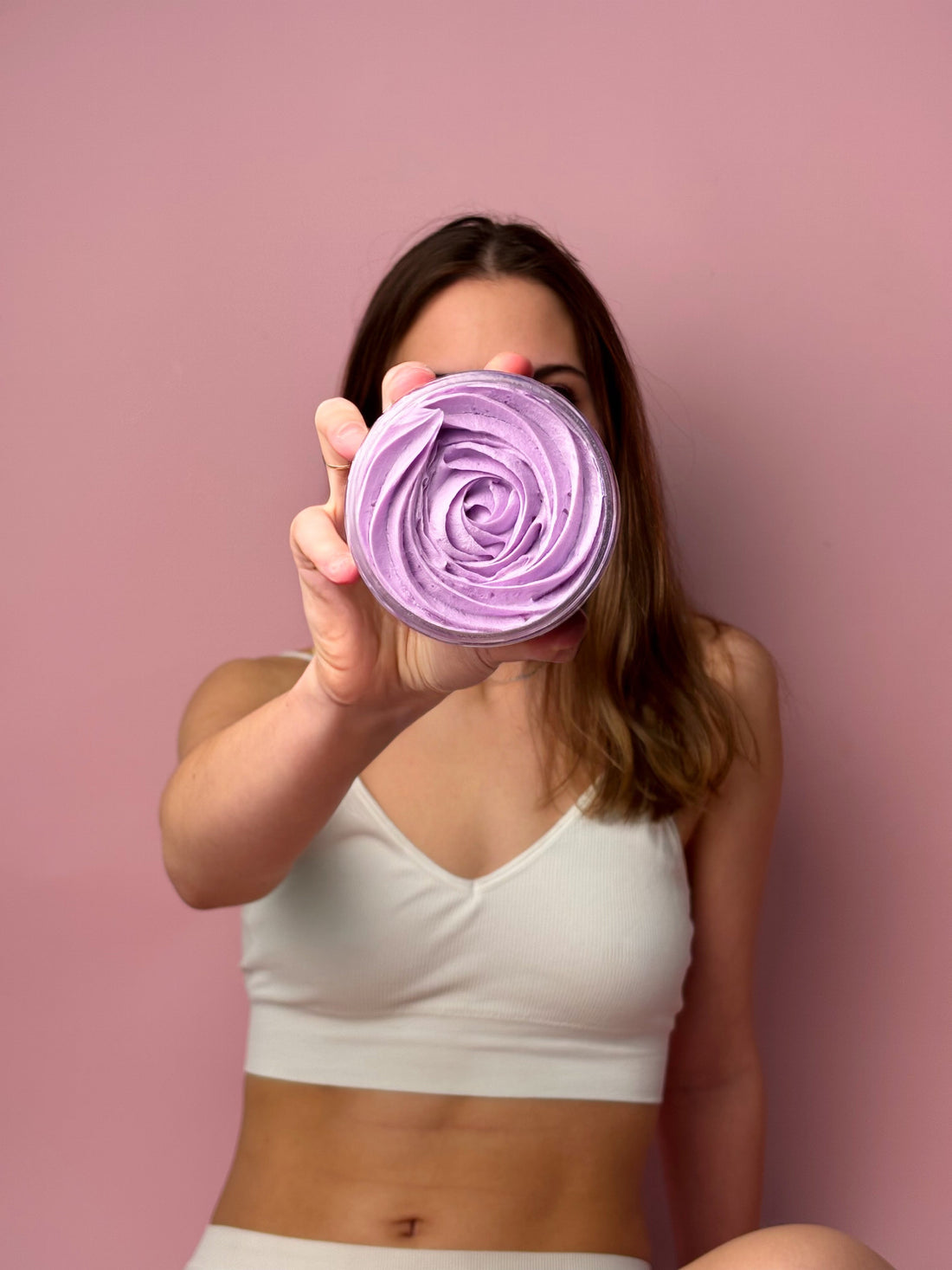 A woman holding a vibrant purple jar of whipped vegan body butter from the High Priestess collection against a pink background.