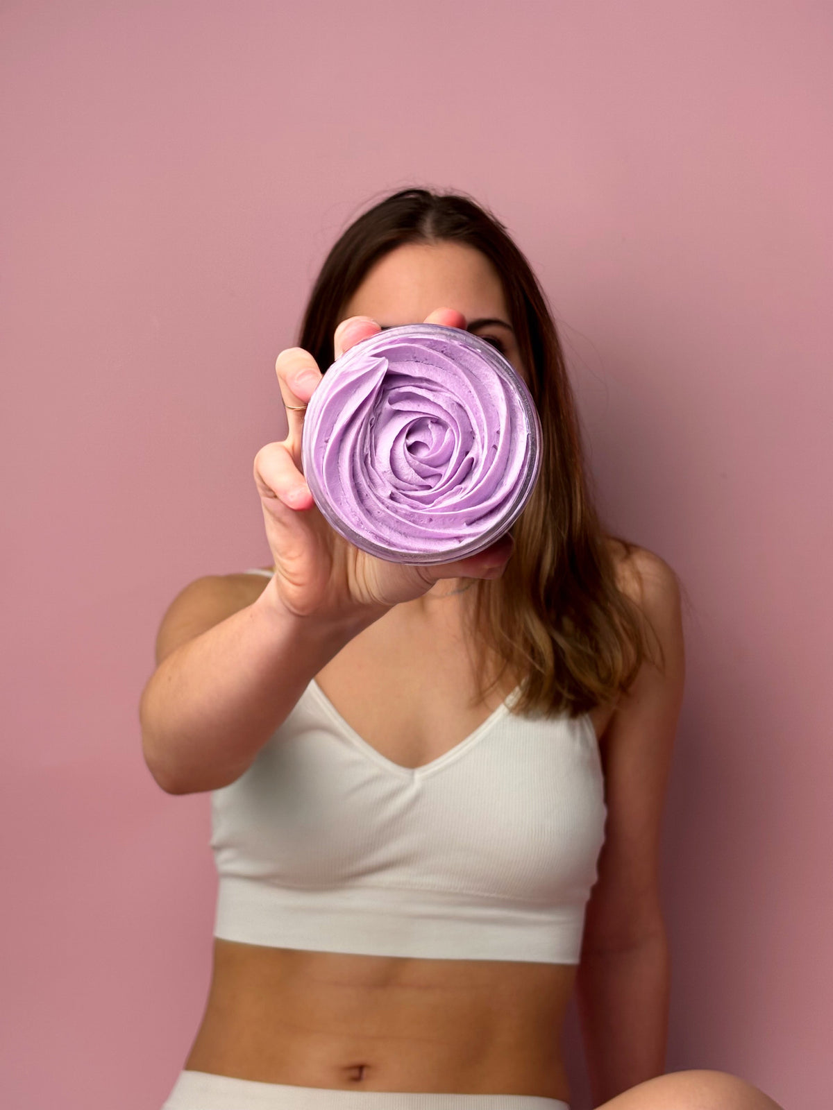 A woman holding a vibrant purple jar of whipped vegan body butter from the High Priestess collection against a pink background.