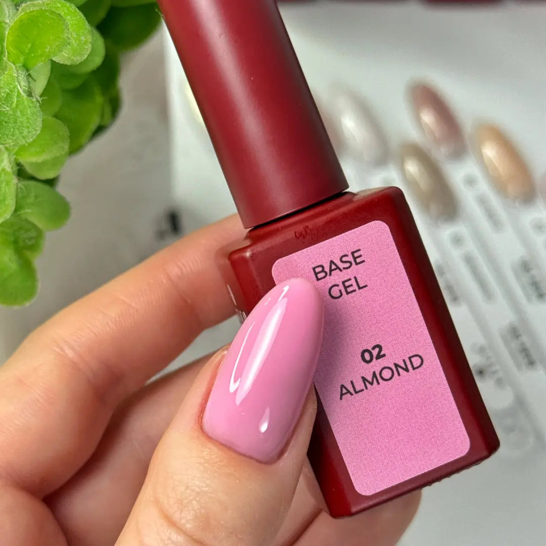 Base gel bottle labeled '02 Almond' held by a hand with pink nail polish, against a blurred background.