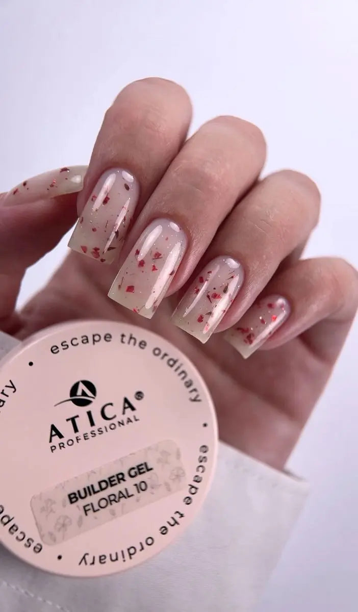A close-up of a hand with nails painted in a sheer nude-pink gel with small red glitter flecks, holding a round container labeled ATICA Professional Builder Gel Floral 10.