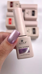 A hand with a metallic lavender-purple manicure holds an ATICA Professional nail system bottle labeled '010 Satin'. In the blurred background, several other ATICA nail polish bottles are stacked on a white surface.