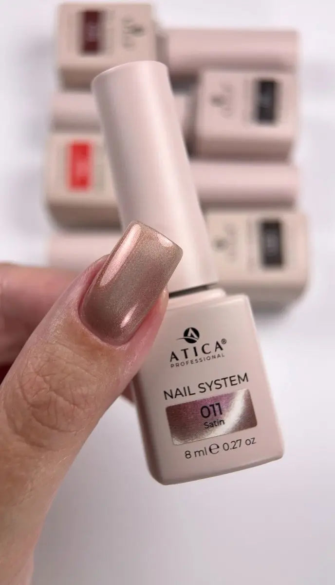 A hand holding an 8ml bottle of ATICA Professional Nail System shade 011 Satin, showing a metallic champagne-gold nail polish swatch on the fingernail. Other bottles of the nail system are blurred in the background.