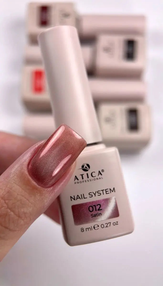 A hand holding a bottle of ATICA Professional Nail System gel nail polish in shade 012 Satin. The nail polish has a metallic rose gold shimmer, and several other bottles are blurred in the background.