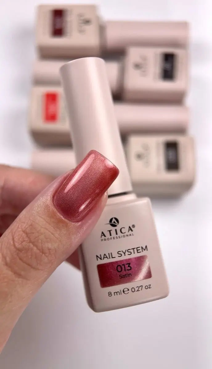 A hand holding an Atica Professional nail polish bottle with a metallic rose pink shade, with other bottles blurred in the background.