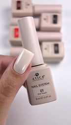 A hand holding a bottle of ATICA Professional Nail System shade 01 One Touch, showing a glossy off-white manicured fingernail, with blurred matching nail polish boxes in the background.