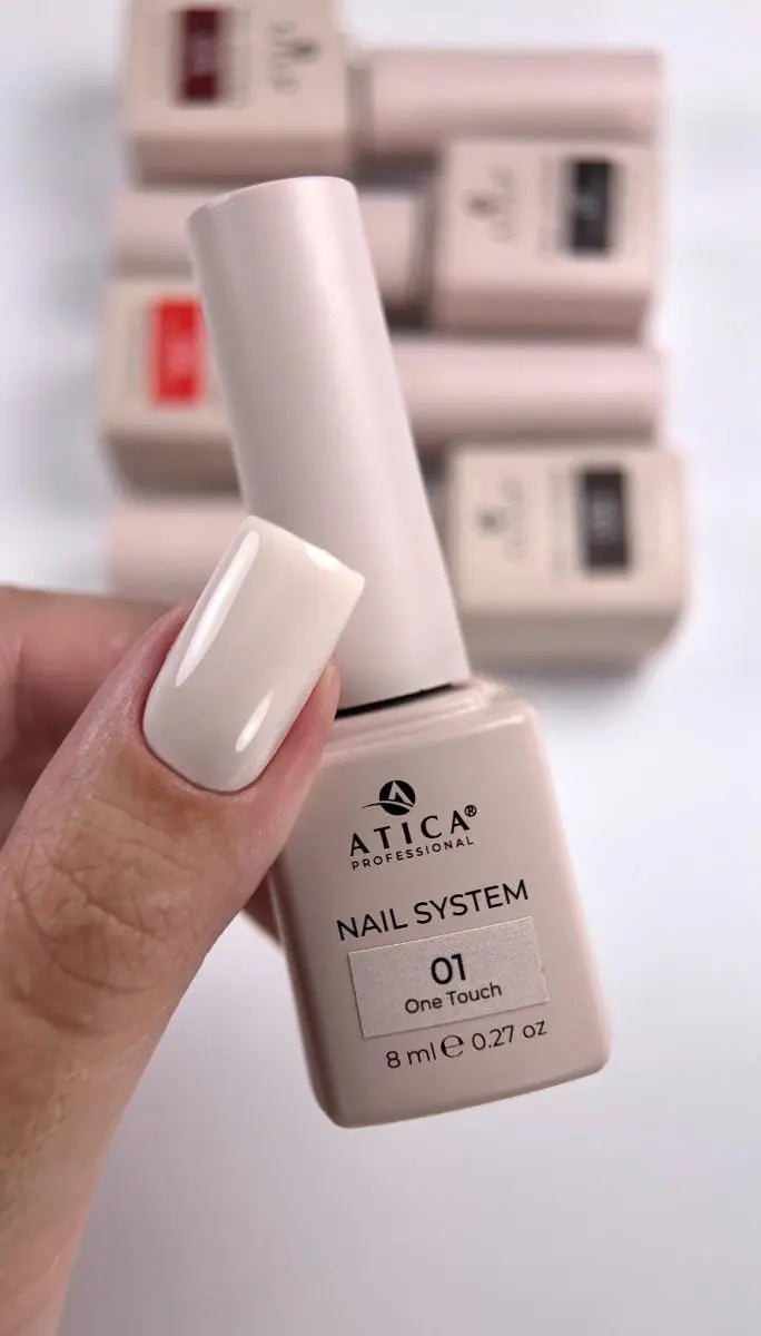 A hand holding a bottle of ATICA Professional Nail System shade 01 One Touch, showing a glossy off-white manicured fingernail, with blurred matching nail polish boxes in the background.