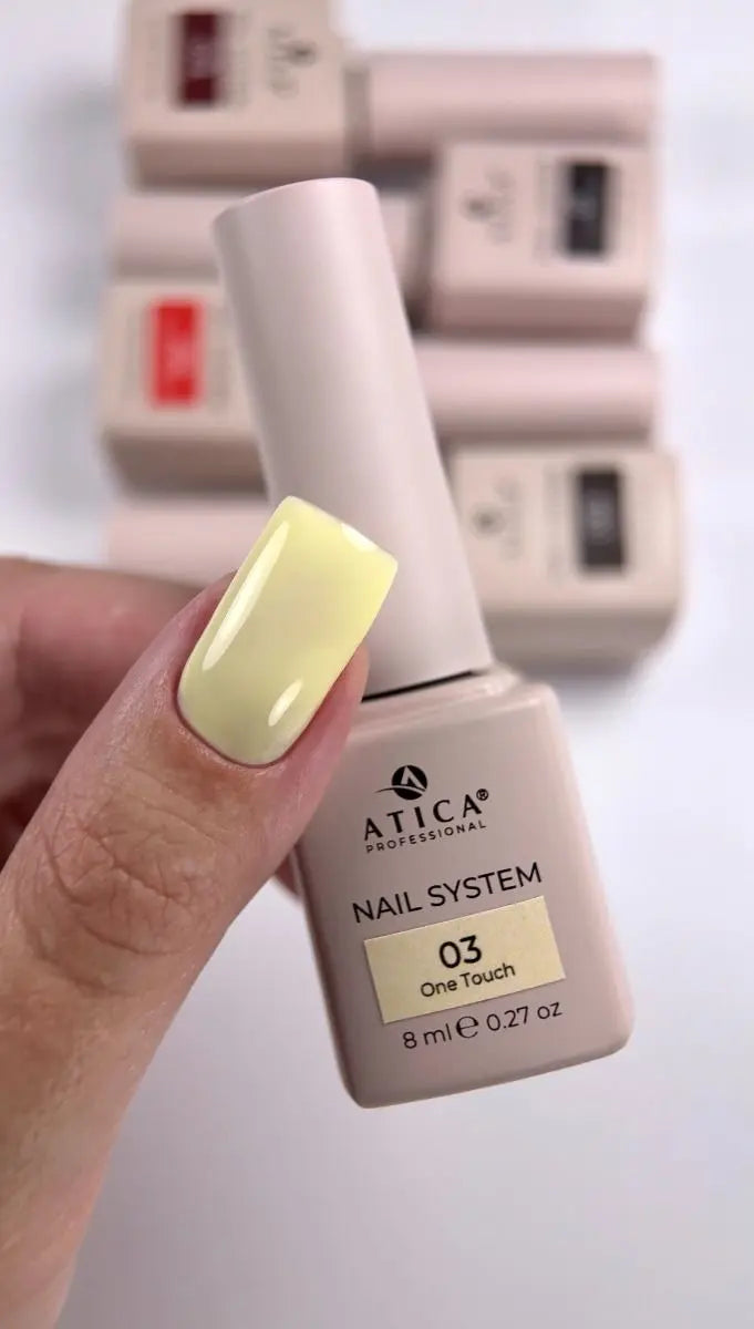 A hand with a pale yellow manicure holds an ATICA Professional nail polish bottle labeled shade 03 One Touch. The bottle is a light beige/taupe color, and several other bottles are stacked in a blurred background.