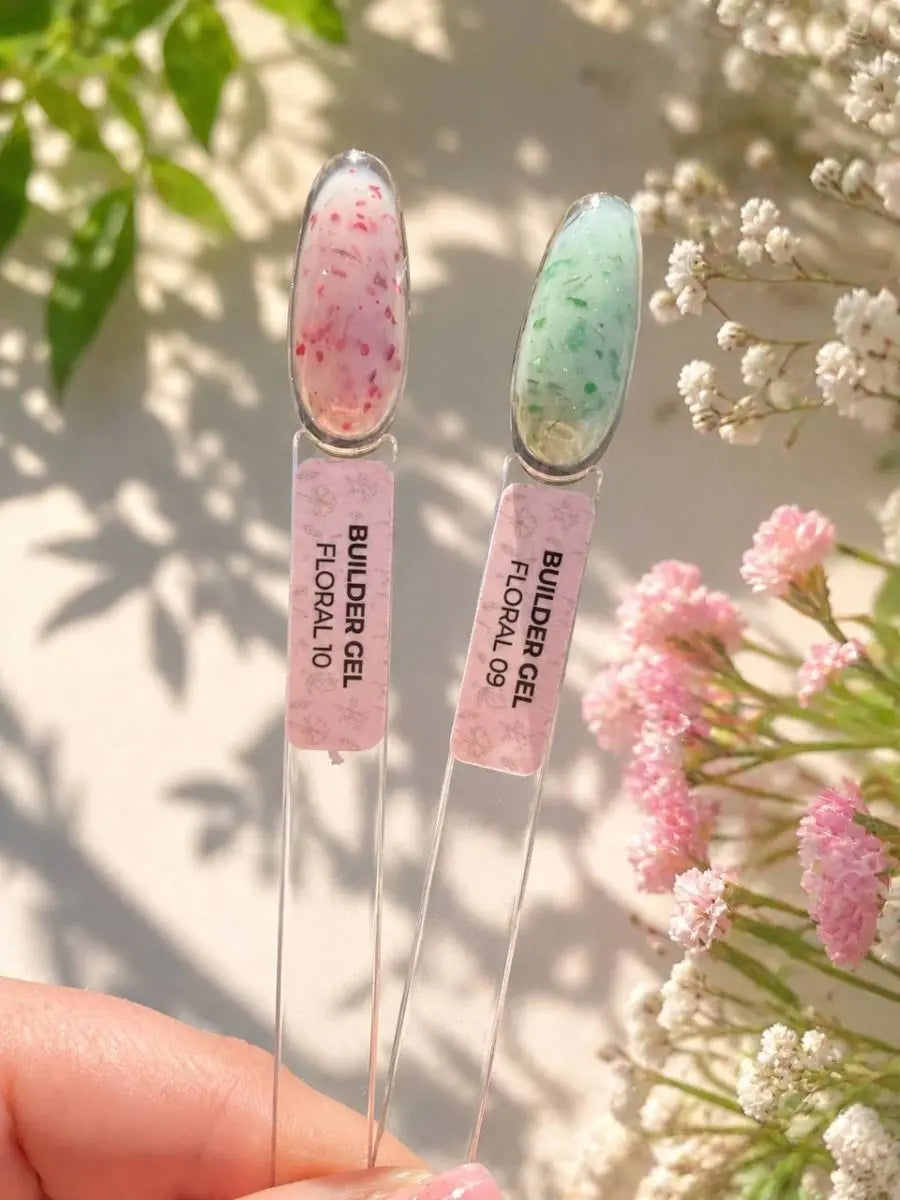 Two nail art gel swatches held in a hand, labeled 'BUILDER GEL FLORAL 10' and 'BUILDER GEL FLORAL 09', with pink and mint green floral designs, set against a background of soft pink flowers and green leaves.