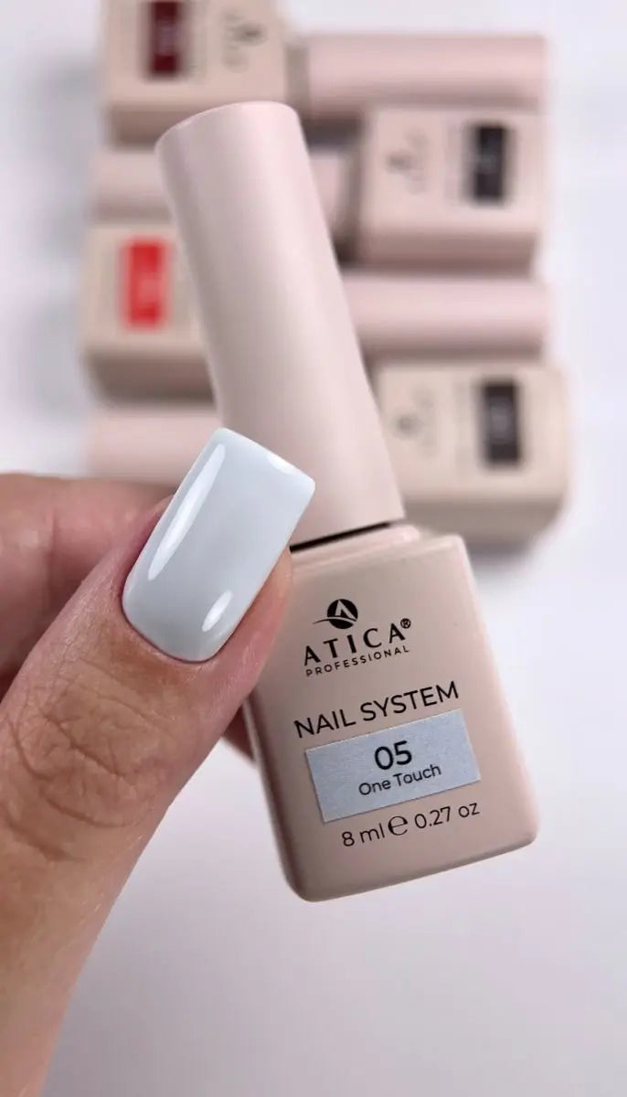 A hand holding a bottle of ATICA Professional Nail System One Touch gel nail polish in shade 05, showing a glossy light blue-gray painted fingernail, with other bottles blurred in the background.