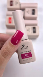 A hand with bright magenta nail polish holds an ATICA Professional nail system bottle labeled shade 07 One Touch. Other ATICA bottles are blurred in the background on a white surface.