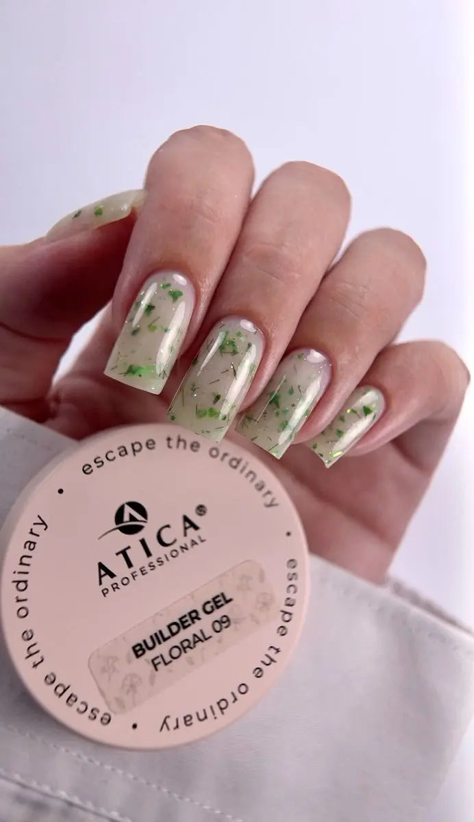 A hand with nails painted in a clear gel with green floral flecks, holding a round pink container of ATICA Professional Builder Gel in Floral 09.