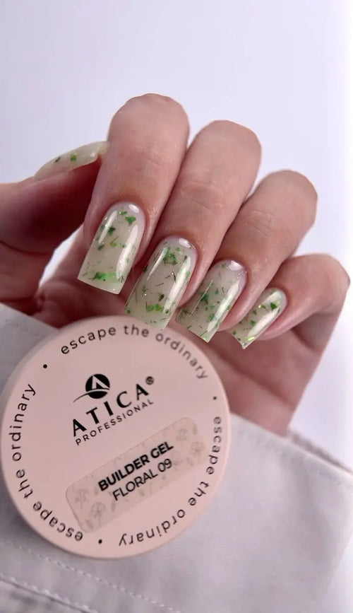 A hand with nails painted in a clear gel with green floral flecks, holding a round pink container of ATICA Professional Builder Gel in Floral 09.
