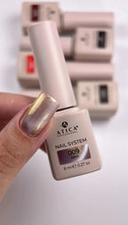 A hand holding a bottle of ATICA Professional Nail System gel nail polish in shade 009 Satin. The nail polish has a shimmering metallic gold finish, and other bottles are blurred in the background.