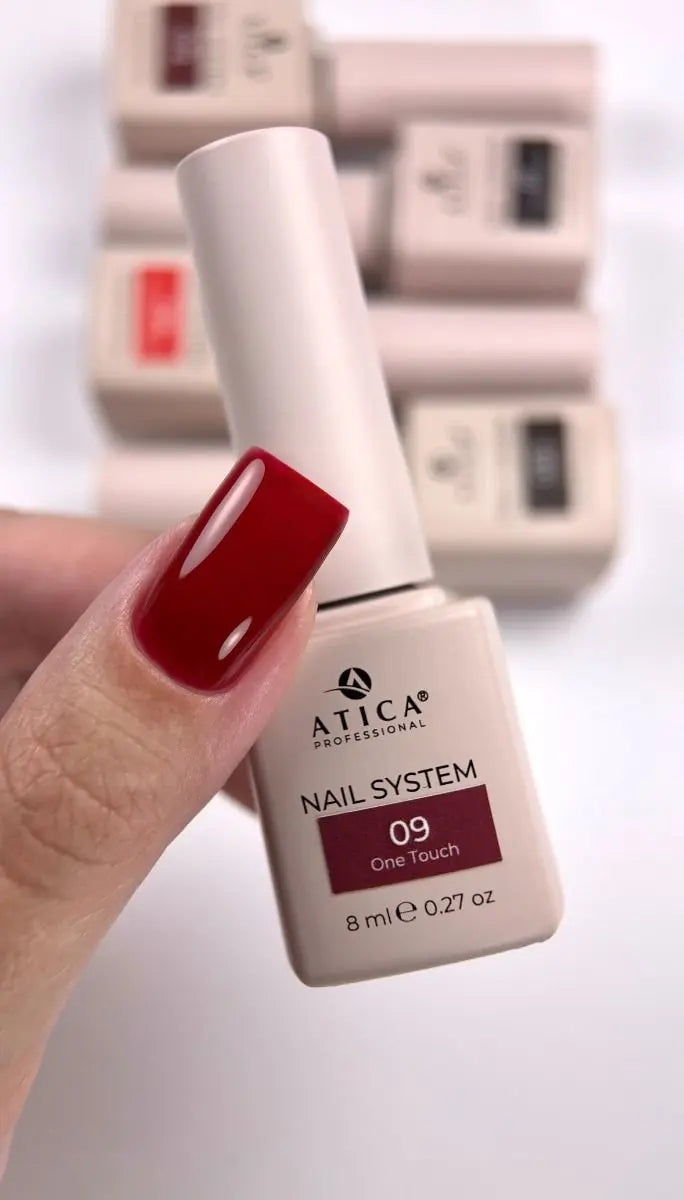 A hand with a glossy deep red manicured fingernail holds an ATICA Professional nail polish bottle labeled 'NAIL SYSTEM 09 Cherry Touch'. In the blurred background, several other ATICA nail polish bottles are stacked on a white surface.
