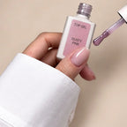 A hand wearing a white long-sleeve shirt holds a bottle of Top Gel nail polish in the shade Dusty Pink, with the applicator brush showing the dusty pink gel polish on a neutral beige background.