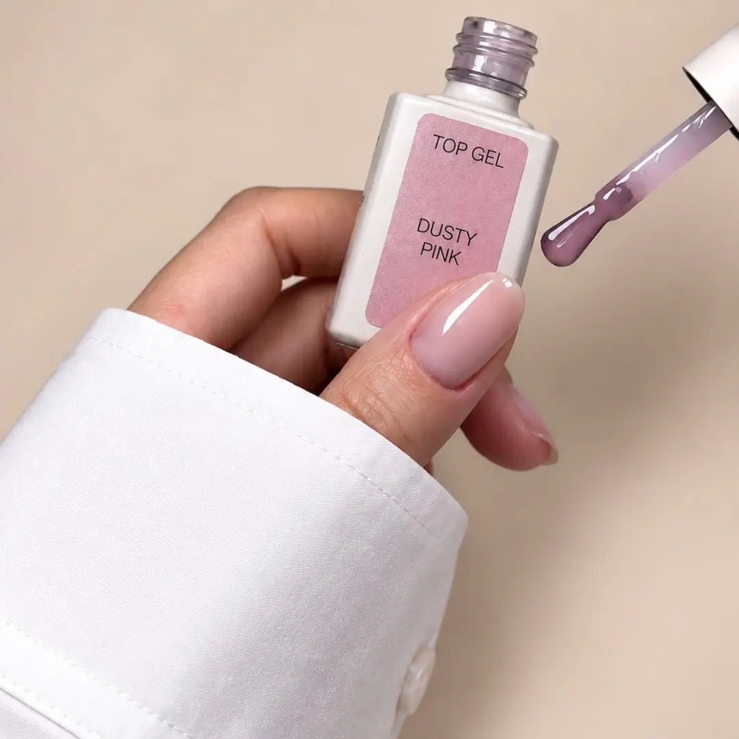 A hand wearing a white long-sleeve shirt holds a bottle of Top Gel nail polish in the shade Dusty Pink, with the applicator brush showing the dusty pink gel polish on a neutral beige background.