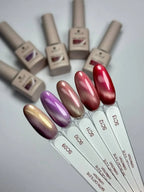 Six nail polish bottles and matching nail swatch sticks arranged on a light gray background, showing different color shades including metallic gold, lavender, champagne, pink, red, and a shimmery purple.