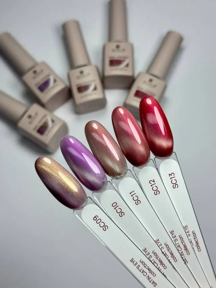 Six nail polish bottles and matching nail swatch sticks arranged on a light gray background, showing different color shades including metallic gold, lavender, champagne, pink, red, and a shimmery purple.