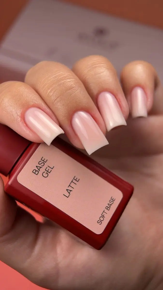 Hand with pink nail polish holding a bottle labeled 'Base Gel Latte' on a blurred background