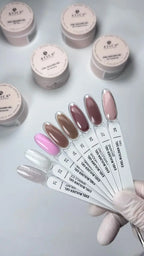 A hand wearing a white glove holds a fan of nail gel color swatch sticks showing various shades, with multiple small jars of ATECA gel polish powder in the background on a light gray surface.