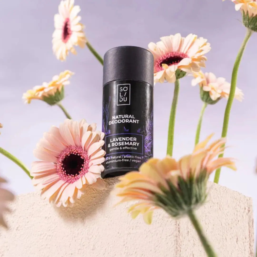 Black deodorant stick with lavender and rosemary scent surrounded by flowers on a light surface.