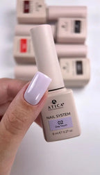 A hand holding an Atica Professional nail polish bottle labeled 'Nail System 02 One Touch' in a pale lavender shade, with other matching bottles blurred in the background.