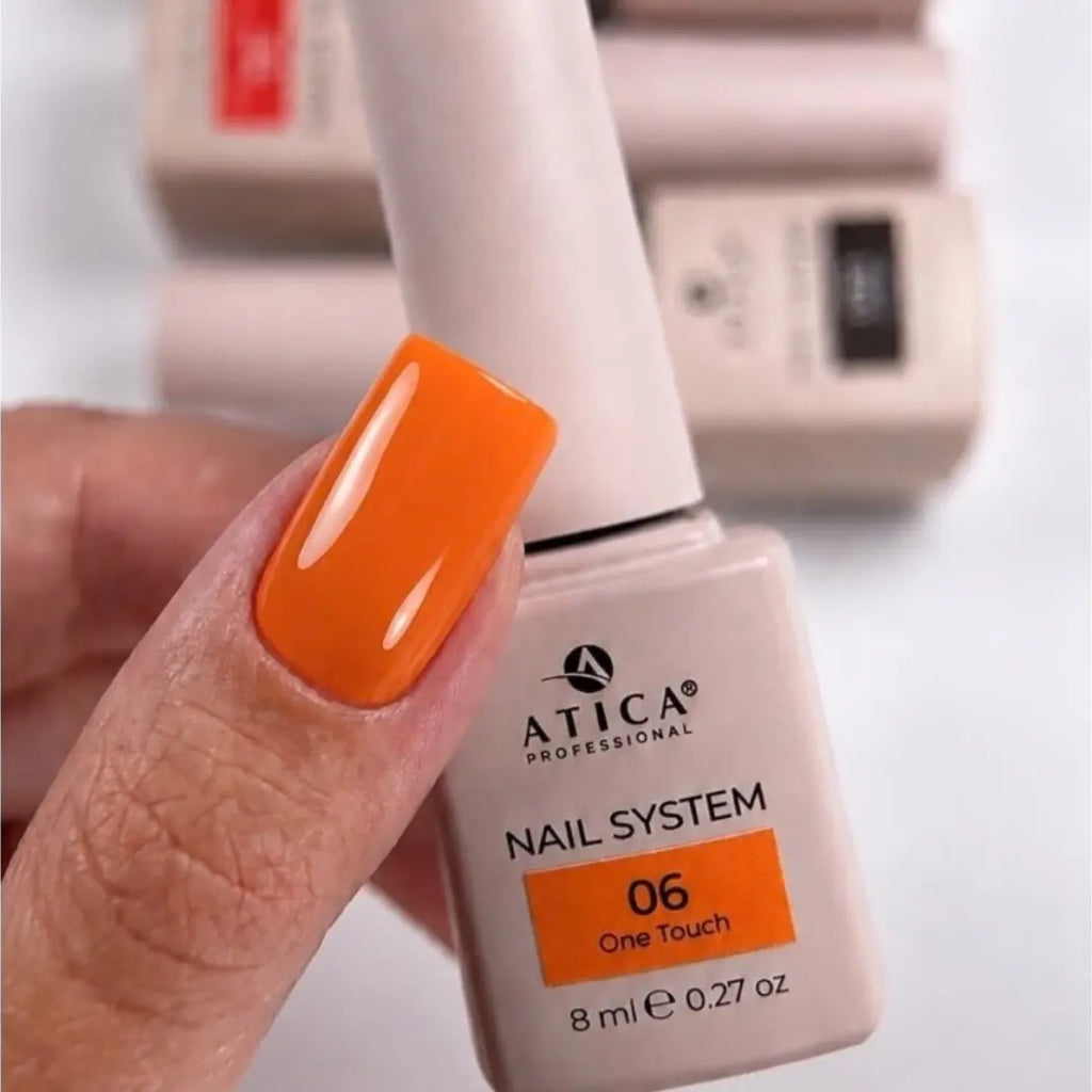 A hand holding an ATICA Professional nail polish bottle showing shade 06 One Touch, with a glossy bright orange nail painted on the fingernail. Other bottles are blurred in the background.