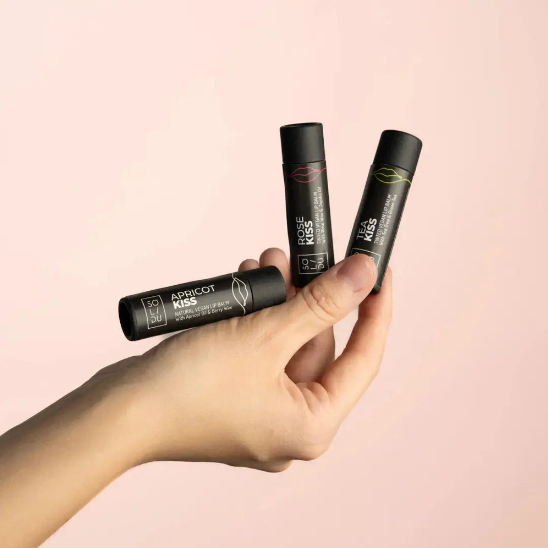 Hand holding three lip balm tubes with visible branding on a pink background