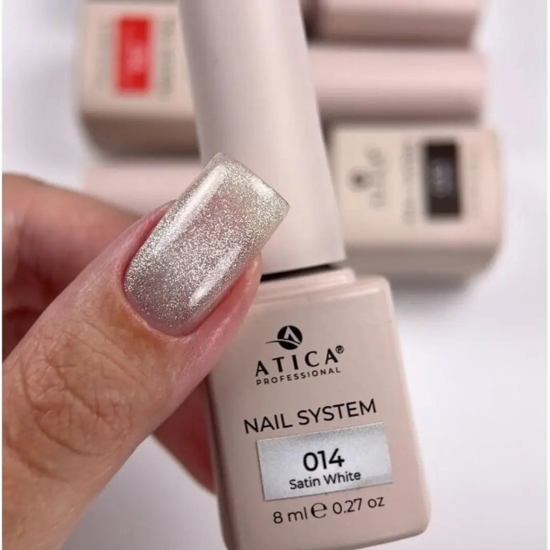 A hand holding an Atica Professional nail system bottle labeled 014 Satin White, showing a glittery silver nail finish on the fingernail, with blurred bottles in the background.