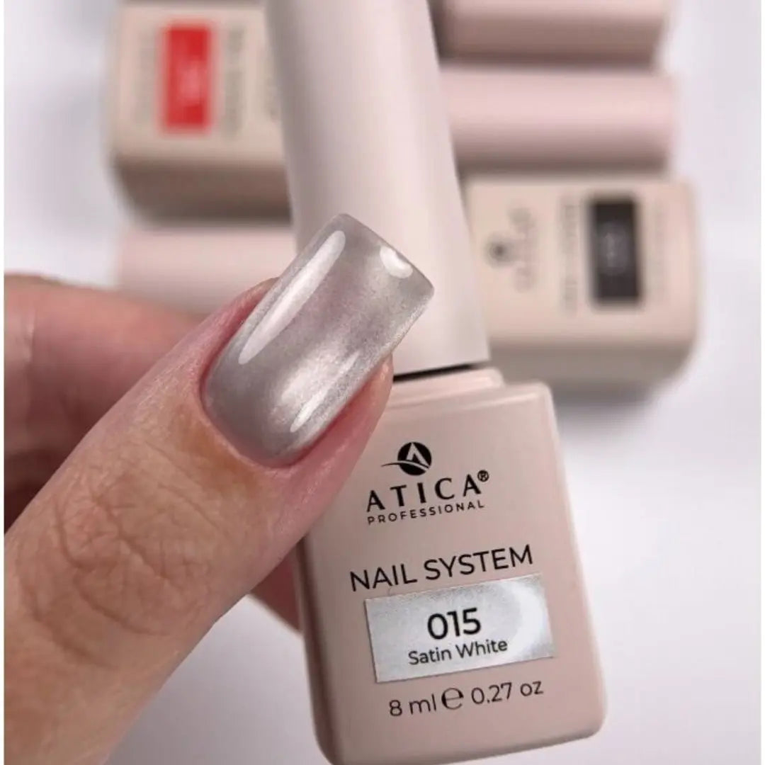 A close-up of a hand with a glossy metallic silver nail polish swatch, holding an ATICA Professional nail system bottle labeled '015 Satin White' against a soft white background with other bottles blurred behind.