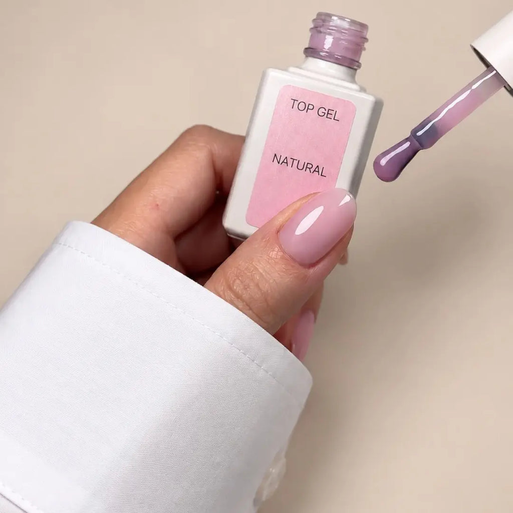 A hand holding a bottle of top gel nail polish labeled 'TOP GEL NATURAL' with a brush applicator showing the glossy light pink polish, against a plain beige background.