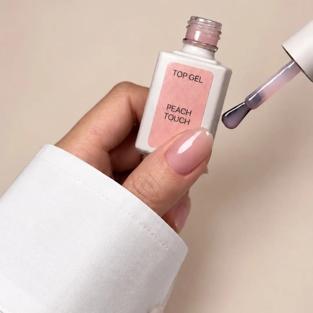 A hand holding a bottle of nail top gel labeled 'TOP GEL PEACH TOUCH' next to a brush applying the pale pink gel to a fingernail, against a soft beige background.