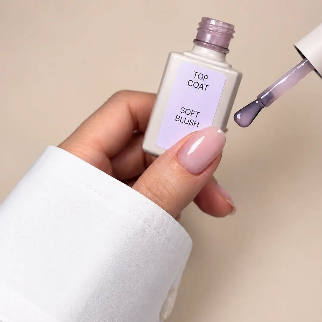 A hand with a pale pink manicure holds a square bottle labeled 'TOP COAT SOFT BLUSH' while a brush applicator with a lavender-purple product extends from the open bottle against a warm beige background.
