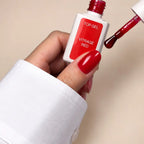 A hand with glossy red nail polish holds a bottle of Top Gel Vitrage Red top coat, with the applicator brush dipped in the red gel against a warm beige background.