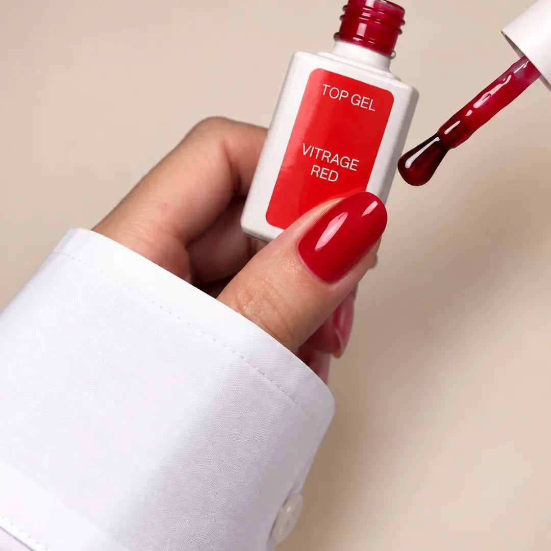 A hand with glossy red nail polish holds a bottle of Top Gel Vitrage Red top coat, with the applicator brush dipped in the red gel against a warm beige background.