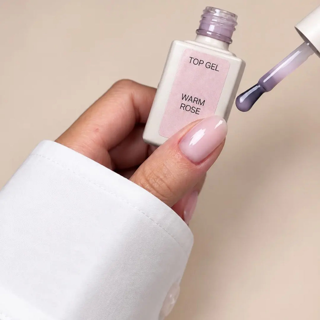 A hand with a glossy pale pink nail polish holds a bottle labeled 'TOP GEL WARM ROSE' next to a brush swiping purple polish onto a beige background.