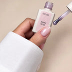 A hand with a glossy pale pink nail polish holds a bottle labeled 'TOP GEL WARM ROSE' next to a brush swiping purple polish onto a beige background.