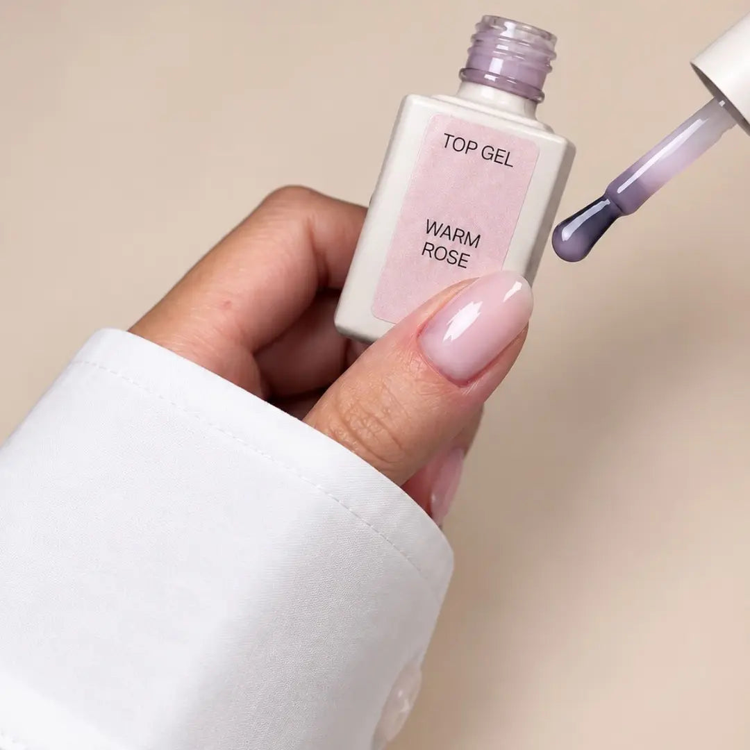 A hand with a glossy pale pink nail polish holds a bottle labeled 'TOP GEL WARM ROSE' next to a brush swiping purple polish onto a beige background.